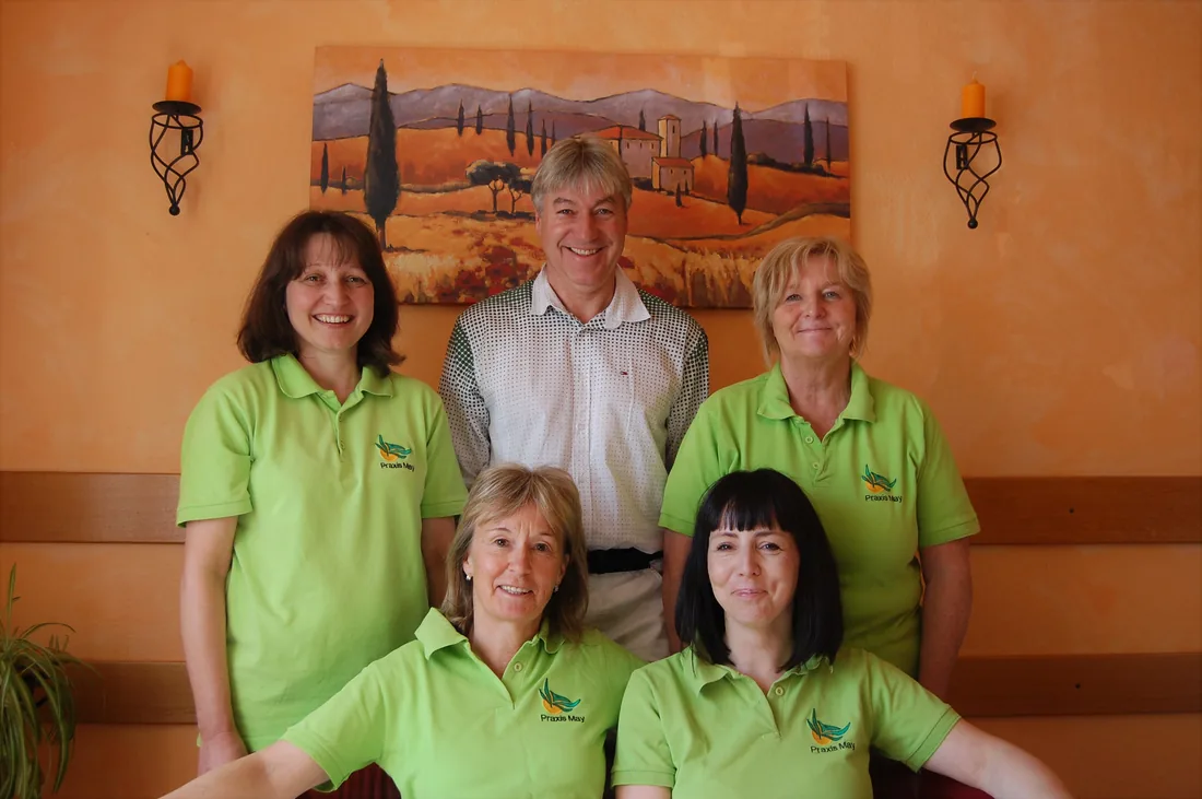 Das Praxis-Team: Hanspeter May steht in der Mitte, um ihn vier Mitarbeiterinnen in grünen Poloshirts mit dem Praxislogo, vor einer orangefarbenen Wand mit einem Toskana-Gemälde.